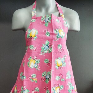 May include: Pink apron with a fruit print featuring pears and cherries. The apron has two pockets.