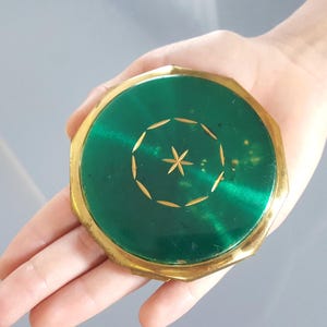Può includere: Uno specchietto compatto rotondo con bordi ottagonali, caratterizzato da una cornice in metallo color oro lucido e una parte superiore in smalto verde smeraldo brillante. Il centro della superficie verde presenta un delicato motivo a stella dorata circondato da un disegno circolare di piccole foglie.