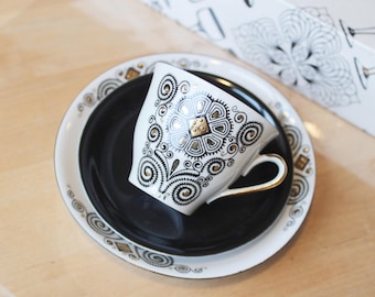 May include: A white ceramic teacup and saucer set featuring intricate black and gold geometric patterns. The teacup has a gold-trimmed handle and a floral-inspired design, while the saucer has a solid black center and a white rim with gold accents.