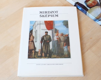 May include: A book titled "Mirdzot Šķepiem" with a painting on the cover depicting a group of people in a village setting. The painting features a man in a military uniform holding a rifle, and a red flag is visible in the background. The text "Veltits Latvijas Sarkanaiem Strelnekiem" is printed below the painting.