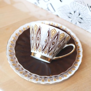May include: A vintage teacup and saucer set. The white teacup features a gold handle and is decorated with brown and gold geometric patterns. The saucer is brown with a gold rim and a white outer edge with gold detailing. The set is on a light wood surface.