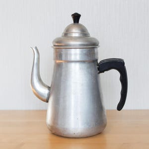 Könnte beinhalten: Eine Vintage-Kaffeekanne aus Aluminium mit schwarzem Griff und Knauf. Die Kanne hat einen gebogenen Ausguss und einen abgerundeten Körper, der sich zum Brühen von Kaffee eignet. Die Kanne steht auf einer Holzoberfläche.