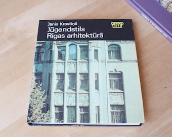 May include: A book titled "Jūgendstils Rīgas arhitektūrā" by Janis Krastins. The cover features a teal and white building facade with many windows. The book is on a wooden surface.