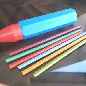May include: A large blue and red plastic pen-shaped object with a black tip, accompanied by six thin, hexagonal-shaped colored pencils in shades of maroon, green, red, light blue, orange, and lime green arranged in a parallel row.
