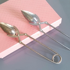 May include: Two silver-toned metal tea infusers with a pointed, perforated oval head and a long, spring-loaded handle featuring a circular loop at the end. The infusers are constructed with a hinged mechanism that allows the two halves to open.