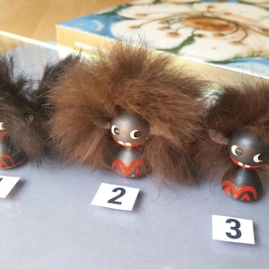 Puede incluir: Tres pequeñas figuras negras y redondeadas con mechones de pelo castaño en la parte superior, ojos blancos y detalles pintados en rojo en la base. Cada figura está etiquetada con una pequeña tarjeta blanca numerada 1, 2 o 3 frente a ella.