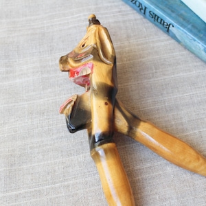May include: A vintage wooden nutcracker in the shape of a horse's head. The nutcracker is primarily light brown with darker brown accents and red detailing in the mouth. The handles are crossed and extend downward.