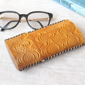 May include: A tan leather eyeglasses case with a detailed embossed pattern and black stitching. Black-framed glasses with gold accents rest beside the case. The case is rectangular and has a textured surface.