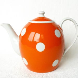 May include: A bright orange ceramic teapot featuring a pattern of large white polka dots. The teapot includes a matching removable lid with a small knob handle, a curved white spout, and a white handle on the side for pouring tea.