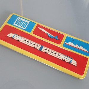 May include: A rectangular metal tin with a gold-coloured rim featuring the word Varia in a blue box. The lid displays a red background with a white passenger train, a white aeroplane, and a white ship with red accents on blue background panels.