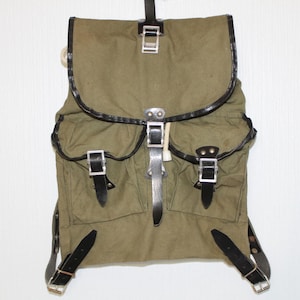 Puede incluir: Una mochila de lona verde oliva con una solapa superior grande, dos bolsillos delanteros y correas de cuero negro con hebillas metálicas plateadas. Los bordes de la solapa y los bolsillos tienen ribetes negros y cuenta con correas ajustables.