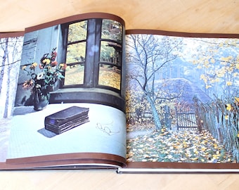 May include: Open book with two colorful images. One shows a vase of flowers by a window, a stack of books, and a table. The other depicts a rural scene with a fence, trees, and a house. The book is open on a light-colored surface.