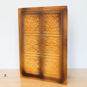Op de afbeelding: Een rechthoekig, amberkleurig decoratief paneel met een herhalend vierkant patroon. Elk vierkant heeft een sierlijk, wervelend ontwerp. Het paneel heeft een textuur en een donkerbruine rand.