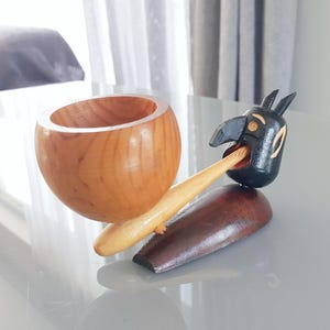 May include: A wooden ornament comprising a light brown bowl and a black bird-like figure with a long, light brown beak. The bird rests on a dark brown base. The bowl is connected to the bird's beak.
