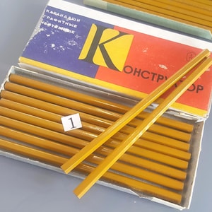 May include: A box of yellow hexagonal graphite pencils with a white label featuring the number 1. The box lid is blue, yellow, and red with Cyrillic text. Two pencils are placed diagonally across the top of the box, while others are inside.