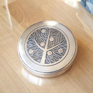 Op de afbeelding: Een ronde, zilverkleurige sieradendoos met een gedetailleerd boomontwerp op het deksel. De boom heeft bladeren en fruit, binnen een cirkelvormige rand. De doos staat op een houten oppervlak.