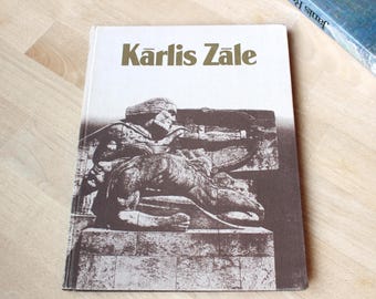 May include: A hardcover book with a white cover featuring the brown text Kārlis Zāle at the top. Below the text is a brown-toned illustration of a stone sculpture depicting a figure with a bow and arrow positioned on a block base.