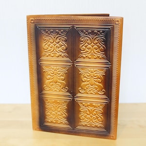 May include: A rectangular notebook with a tan and dark brown cover featuring a repeating embossed pattern of symmetrical scrollwork. The cover has a decorative border with a linear texture and a smooth spine, presenting a uniform grid design.