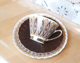 May include: A white porcelain teacup with a brown and gold geometric pattern sits on a dark brown saucer, which is placed atop a larger white plate featuring a matching brown and gold decorative border with a scalloped edge and gold trim.