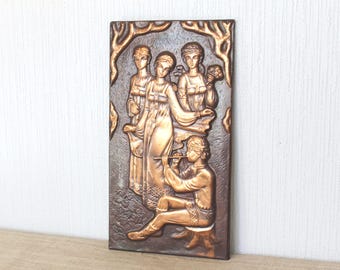 May include: A rectangular copper-colored relief sculpture depicting a scene with figures in flowing garments. The artwork features a musician playing a flute, three women, and decorative tree-like elements. The relief is mounted on a dark brown background.