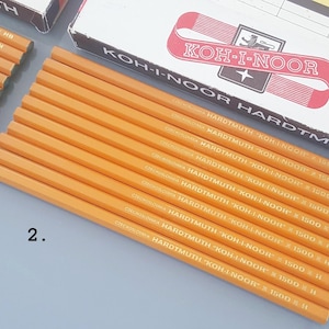 Puede incluir: Una colección de lápices de grafito hexagonales amarillos etiquetados como KOH-I-NOOR HARDTMUTH 1500 H, dispuestos en fila junto a una caja de cartón blanca y roja. El número 2 está impreso en la superficie cerca de los lápices sin punta.