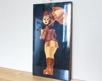 May include: Wooden marquetry art piece depicting a woman holding an umbrella. The artwork features a dark background and a figure in a skirt, jacket, and boots. The umbrella and figure are made of various wood tones.