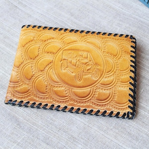 Op de afbeelding: Tan lederen portemonnee met een reliëfontwerp van een klassieke auto. De portemonnee heeft een herhalend cirkelvormig patroon en zwarte stiksels langs de randen. De portemonnee is rechthoekig en lijkt nieuw.