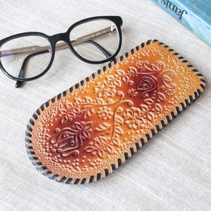 May include: Black eyeglasses rest on a handcrafted, oval-shaped leather case. The case features an embossed floral design in shades of orange and brown, with a black stitched border. A book with a blue cover is partially visible.