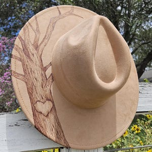 May include: A tan fedora-style hat with a brown tree design and a heart carved into the trunk. The hat is resting on a white fence, with a background of sunflowers and greenery.