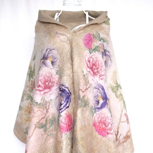 K&ouml;nnte beinhalten: Beigefarbener Kapuzenponcho mit floralem Muster mit rosa und lila Bl&uuml;ten. Der Poncho hat einen Goldschimmer und eine weiche Textur. Das Design umfasst eine Kapuze und eine drapierte Silhouette.