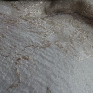K&ouml;nnte beinhalten: Nahaufnahme eines weichen, strukturierten Stoffes in Grau- und Beiget&ouml;nen. Zarte, hellbraune F&auml;den bilden ein subtiles, flie&szlig;endes Muster auf der Oberfl&auml;che. Der Stoff scheint drapiert zu sein, mit sanften Falten und Schatten.