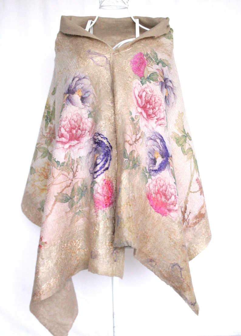K&ouml;nnte beinhalten: Beigefarbener Kapuzenponcho mit floralem Muster mit rosa und lila Bl&uuml;ten. Der Poncho hat einen Goldschimmer und eine weiche Textur. Das Design umfasst eine Kapuze und eine drapierte Silhouette.