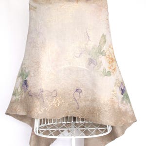 K&ouml;nnte beinhalten: Ein beigefarbener Filzschal mit floralen und abstrakten Designs. Der Schal wird auf einer wei&szlig;en Schaufensterpuppe pr&auml;sentiert. Das florale Design umfasst violette, gr&uuml;ne und orange Farbt&ouml;ne. Die Kanten des Schals haben einen Goldschimmer.