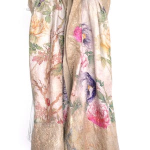 K&ouml;nnte beinhalten: Ein beiger Schal mit einem Blumenmuster mit rosa, lila und gelben Blumen. Der Schal hat einen goldfarbenen Rand und ist &uuml;ber einem wei&szlig;en Kleiderb&uuml;gel drapiert. Das Design des Schals umfasst Zweige und Bl&auml;tter.