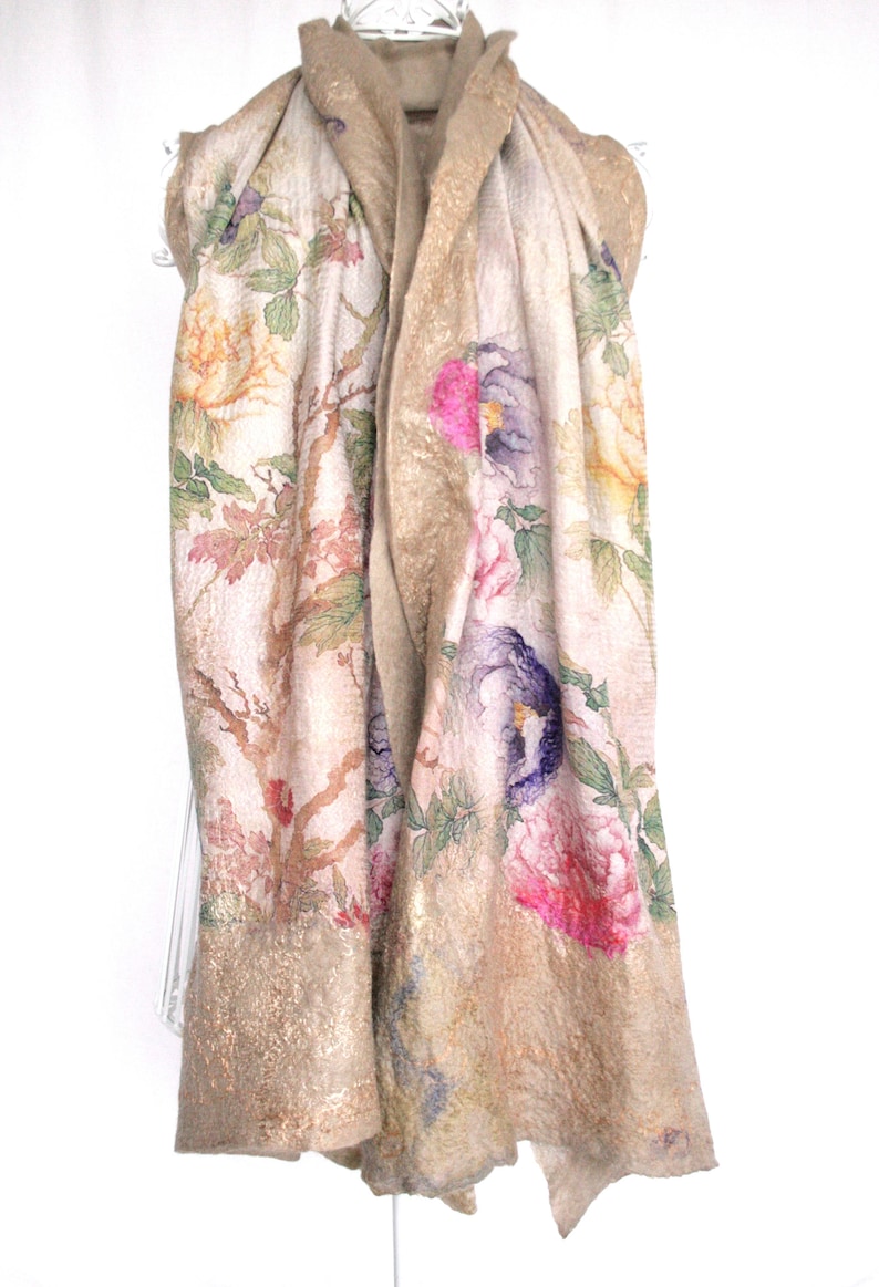 K&ouml;nnte beinhalten: Ein beiger Schal mit einem Blumenmuster mit rosa, lila und gelben Blumen. Der Schal hat einen goldfarbenen Rand und ist &uuml;ber einem wei&szlig;en Kleiderb&uuml;gel drapiert. Das Design des Schals umfasst Zweige und Bl&auml;tter.
