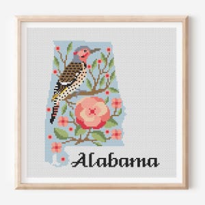 May include: A framed cross-stitch pattern featuring the state of Alabama in light blue, decorated with a northern flicker bird, a camellia flower, and green branches with red berries. The word Alabama is stitched in black at the bottom.