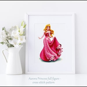 May include: A cross stitch pattern featuring Aurora, the princess from Sleeping Beauty, in a pink gown with a sparkling design. The pattern is framed and displayed on a white surface.