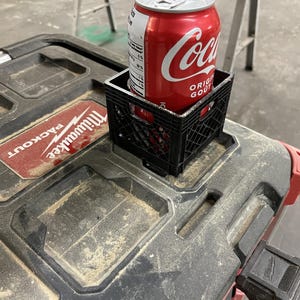 May include: A red Coca-Cola can in a small black plastic crate, resting on a dusty black and red Milwaukee Packout toolbox. The can is partially visible, displaying the Coca-Cola logo and nutritional information.