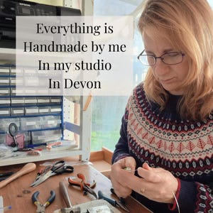 May include: A person wearing a navy blue, red, and white patterned sweater working at a wooden desk with various jewelry-making tools including pliers and a hammer. Text overlay reads: Everything is Handmade by me In my studio In Devon.