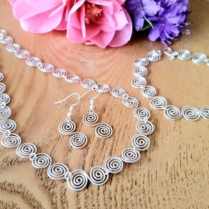 May include: A silver necklace, bracelet and earrings set with a spiral design. The jewellery is displayed on a wooden surface with pink and purple flowers.
