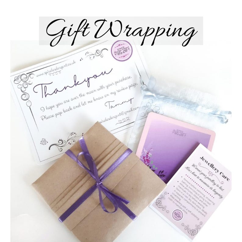 May include: A brown paper gift package tied with a purple ribbon, accompanied by a white thank you card, a small light blue drawstring pouch, a purple card with floral accents, and a white jewellery care instruction card with black text.
