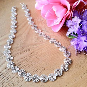 May include: A silver-toned necklace composed of a series of interconnected circular spiral links. The metal chain features a repeating pattern of individual wire coils linked together to form a continuous, flexible strand of metallic silver spirals.