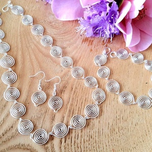 Può includere: Un set di gioielli color argento composto da una collana, un bracciale e un paio di orecchini pendenti. Ogni pezzo è realizzato con spirali di filo metallico interconnesse, caratterizzate da un motivo a bobina circolare costante.