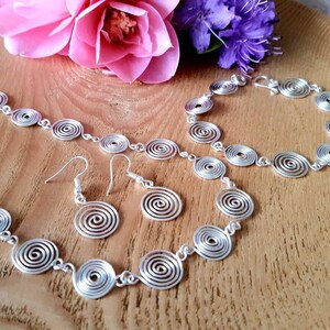 May include: A silver necklace, bracelet and earrings set with spiral-shaped links. The jewellery is displayed on a wooden surface with pink and purple flowers.