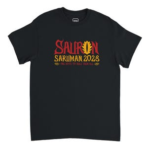 May include: Black t-shirt with the text "SAURON SARUMAN 2028" in red and yellow lettering. The shirt also includes the phrase "ONE VOTE TO RULE THEM ALL" in yellow. The design is centered on the front of the shirt.