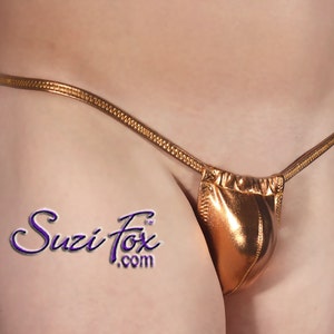 Puede incluir: Un tanga dorado metálico con una pequeña bolsa en la parte delantera. El tanga está hecho de una tela brillante y lisa. El nombre de la marca "Suzi Fox" está impreso en el muslo izquierdo.