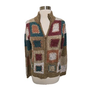 May include: A zip-up sweater featuring a patchwork design of squares in shades of tan, teal, burgundy, and white. The knit garment has a high collar, long sleeves, and a full-length front zipper closure, showcasing a textured, multi-colored pattern.