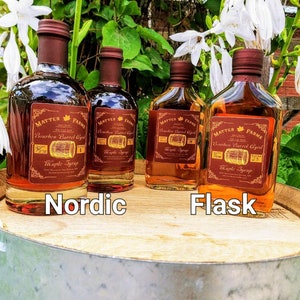 May include: Four glass bottles of maple syrup, labeled "Matter Farms Bourbon Barrel Aged Maple Syrup". The bottles are arranged in a row on a wooden barrel, with the words "Nordic" and "Flask" printed below them.