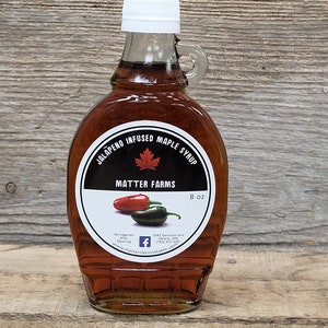 May include: A glass bottle of jalapeno infused maple syrup with a white cap and a black label that says "Jalapeno Infused Maple Syrup" and "Matter Farms". The label also includes a red maple leaf, two jalapenos, and the text "8 oz".