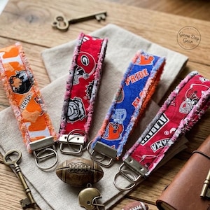 May include: Four fabric wristlet keychains with frayed edges, each featuring distinct college sports team patterns in orange, red, and blue. The wristlets include silver-toned metal hardware with attached split rings for keys.
